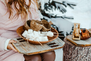 Winter Picnic. Girl's hands with wooden utensils and marshmallows on sticks against a backdrop of snow.