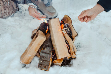 Winter Picnic. Bonfire in the snow and hands with marmalade and sausage on sticks. Horizontal image.