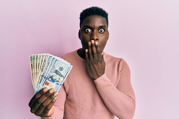 Handsome black man holding dollars covering mouth with hand, shocked and afraid for mistake....