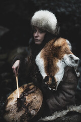 Portrait of a woman wearing fur hat and fox fur pelt on her shoulders.Playing shamanic drum in the forest.