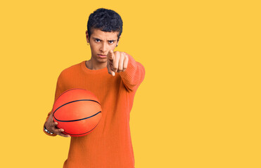 Young african amercian man holding basketball ball pointing with finger to the camera and to you, confident gesture looking serious