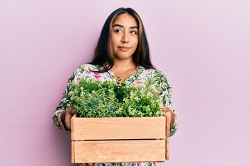 Young latin woman holding wooden plant pot smiling looking to the side and staring away thinking.