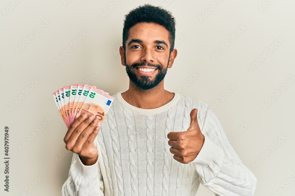 Wall mural Handsome hispanic man with beard holding bunch of 10 euro banknotes smiling happy and positive, thumb up doing excellent and approval sign