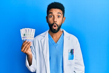 Handsome hispanic man with beard wearing doctor uniform holding 50 colombian pesos scared and amazed with open mouth for surprise, disbelief face