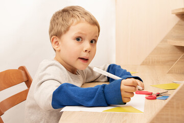 Funny boy with surprised face sit at the table,while drawing or writing.Home education concept or development activities.Online learning.