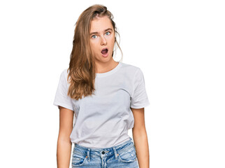 Beautiful young blonde woman wearing casual white t shirt in shock face, looking skeptical and sarcastic, surprised with open mouth