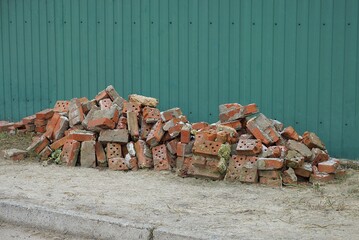 two heaps of pieces of red old bricks on gray ground on the street near the green wall of the fence