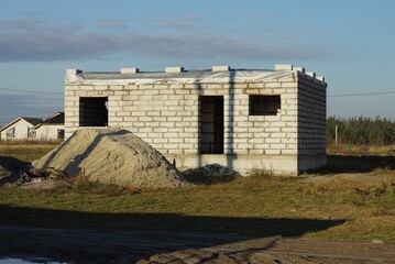 one white private unfinished brick house on a construction site in green grass with a pile of gray sand against a blue sky