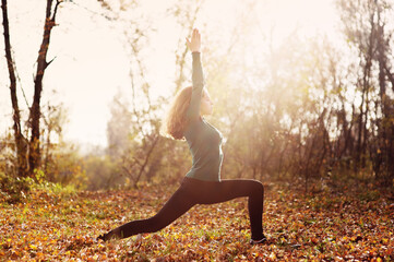 Young caucasian woman with brown hair practicing yoga in the autumn forest. View of a warrior pose.