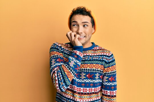Young handsome man wearing wool hat and colorful sweater looking stressed and nervous with hands on mouth biting nails. anxiety problem.