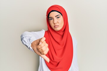 Young beautiful hispanic girl wearing traditional islamic hijab scarf looking unhappy and angry showing rejection and negative with thumbs down gesture. bad expression.