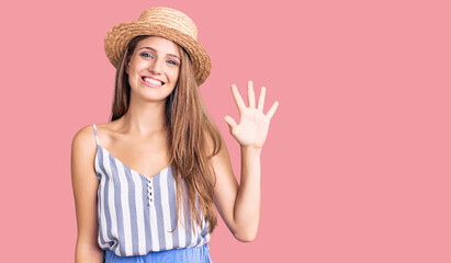 Young beautiful blonde woman wearing summer hat showing and pointing up with fingers number five while smiling confident and happy.