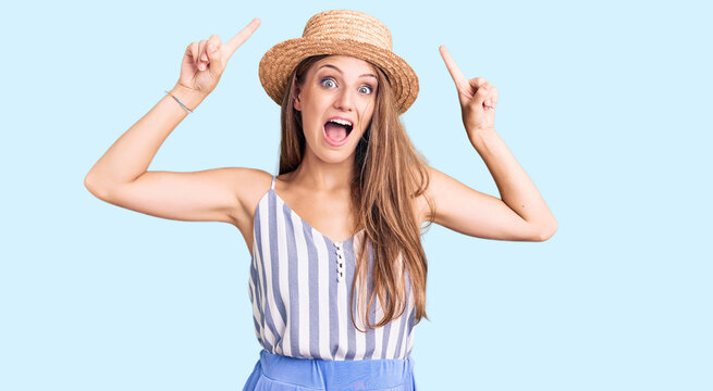 Young beautiful blonde woman wearing summer hat smiling amazed and surprised and pointing up with fingers and raised arms.
