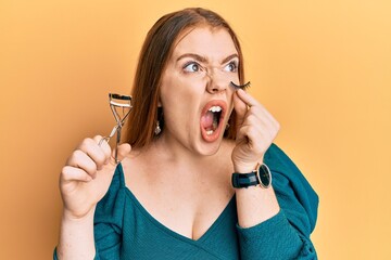 Young beautiful redhead woman holding eyelash curler and fake lashes angry and mad screaming...