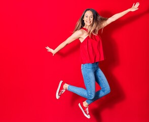 Young beautiful woman wearing casual clothes smiling happy. Jumping with smile on face and arms opened over isolated red background