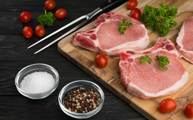 Meat steaks. Raw pork or beef ribeye meat with bone. Ingredients for barbecue. Cherry tomatoes, colored pepper, sea salt and parsley on black wooden background. Veal chunk. Food photography.
