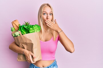 Young blonde girl holding paper bag with bread and groceries covering mouth with hand, shocked and...