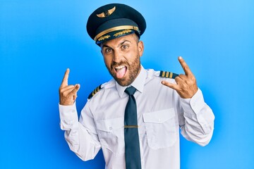 Handsome man with beard wearing airplane pilot uniform shouting with crazy expression doing rock...