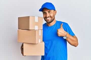 Handsome man with beard wearing courier uniform holding delivery packages doing happy thumbs up gesture with hand. approving expression looking at the camera showing success.