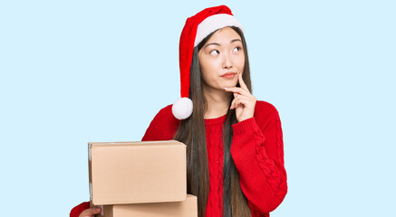 Young chinese woman wearing christmas hat holding delivery package serious face thinking about question with hand on chin, thoughtful about confusing idea