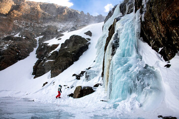 View of a woman standing in front of a frozen waterfall during the winter in Rocky Mountain National Park in Colorado on the Glacier Gorge Trail. Snow, rocks, ice, and mountains can be seen.