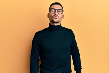 Handsome man with tattoos wearing turtleneck sweater and glasses looking at the camera blowing a...
