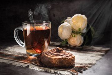 Tea is fragrant black, in a transparent mug, on a light brown napkin, with a snail bun with cinnamon and flowers in the background on a gray background