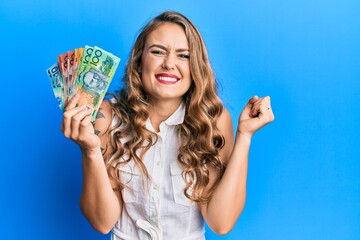 Young blonde girl holding australian dollars screaming proud, celebrating victory and success very...