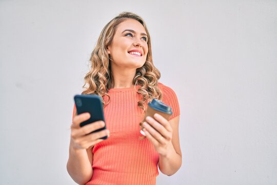 Young blonde girl smiling happy using smartphone and drinking take away coffee at the city.
