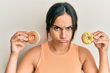 Young brunette woman holding tasty colorful doughnuts skeptic and nervous, frowning upset because...