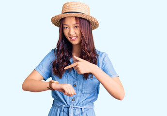 Young beautiful chinese girl wearing summer hat in hurry pointing to watch time, impatience, upset...