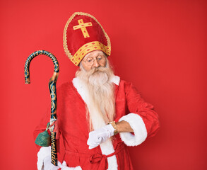 Old senior man with grey hair and long beard wearing traditional saint nicholas costume checking the time on wrist watch, relaxed and confident