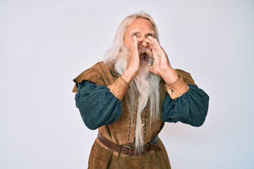 Old senior man with grey hair and long beard wearing viking traditional costume shouting angry out loud with hands over mouth
