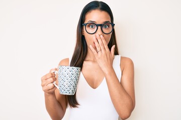 Young beautiful latin girl wearing glasses holding coffee covering mouth with hand, shocked and...