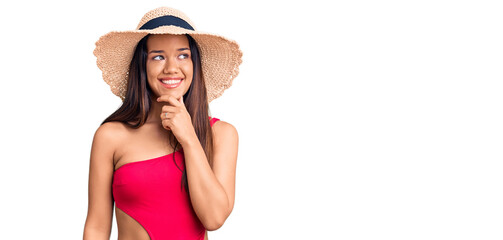 Young beautiful latin girl wearing swimwear and summer hat with hand on chin thinking about question, pensive expression. smiling with thoughtful face. doubt concept.