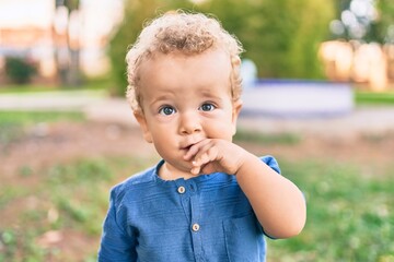Sad little boy putting fingers on mouth touching gums because toothache at the park on a sunny day. Beautiful blonde hair male toddler in pain for new baby teeth outdoors