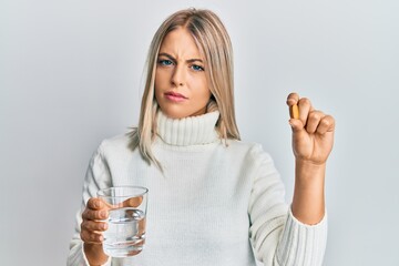 Beautiful blonde woman holding pill and glass of water clueless and confused expression. doubt...