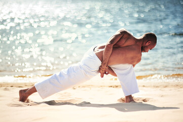 Young caucasian male practicing yoga in white pants on a sandy shore.