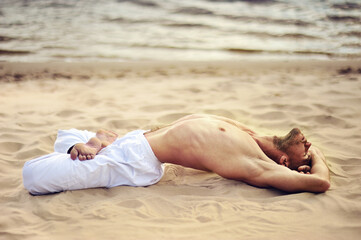 Young caucasian male practicing yoga in white pants on a sandy shore.