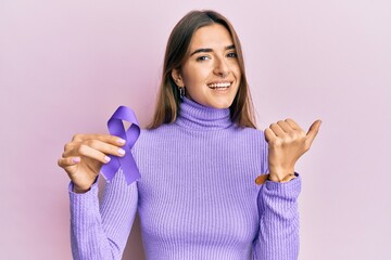 Young hispanic woman holding purple ribbon awareness pointing thumb up to the side smiling happy...