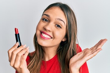 Young brunette woman holding red lipstick celebrating achievement with happy smile and winner expression with raised hand