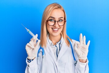 Beautiful caucasian woman wearing doctor stethoscope holding syringe doing ok sign with fingers, smiling friendly gesturing excellent symbol