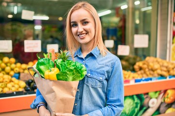 Young blonde girl smiling happy holding groceries paper bag standing at the supermarket