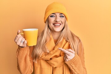 Obraz na płótnie Canvas Young caucasian woman wearing winter scarf and drinking a cup of hot coffee smiling happy pointing with hand and finger