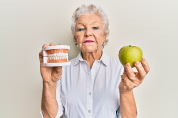 Senior grey-haired woman holding green apple and denture teeth relaxed with serious expression on...