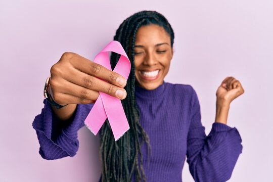 African american woman holding pink cancer ribbon screaming proud, celebrating victory and success very excited with raised arm