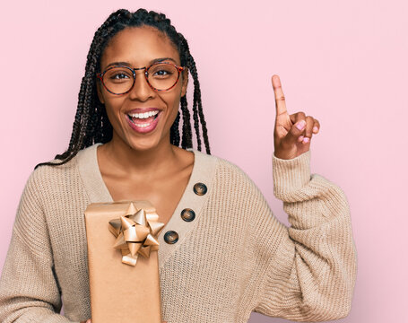 African american woman holding gift surprised with an idea or question pointing finger with happy face, number one