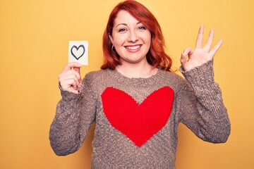 Young beautiful redhead woman holding reminder with heart over isolated yellow background doing ok sign with fingers, smiling friendly gesturing excellent symbol