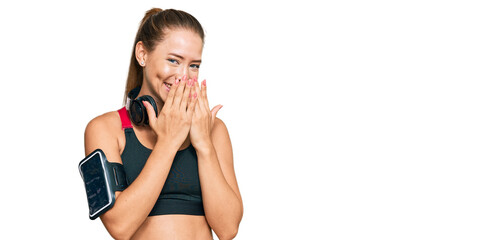 Beautiful blonde woman wearing gym clothes and using headphones laughing and embarrassed giggle...