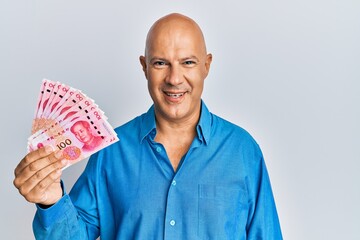 Middle age bald man holding 100 yuan chinese banknotes looking positive and happy standing and smiling with a confident smile showing teeth